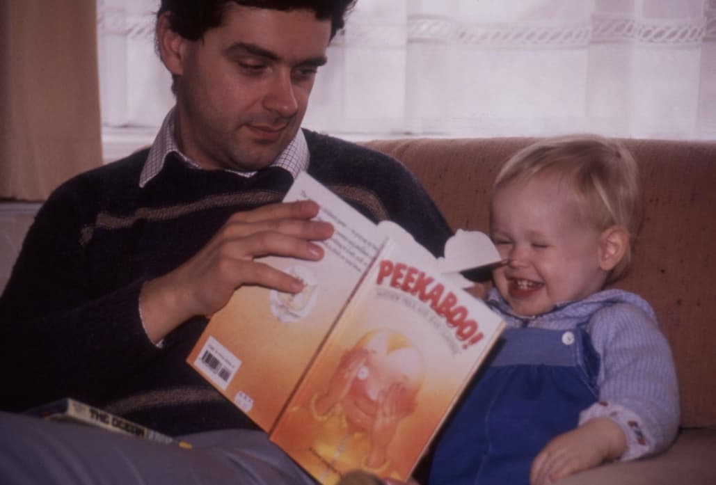 An old photograph of baby Pete, with his father. His dad is holding a book titled 'Peekaboo'. Baby pete is laughing.